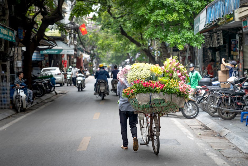 A street scene featuring a flower vendor pushing a bicycle loaded with a vibrant bouquet of fresh flowers, including pink lilies, white hydrangeas, yellow chrysanthemums, and assorted greenery, wrappe